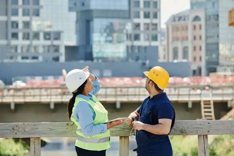 Two engineers in hard hats and vests review plans on a tablet beside a city construction site