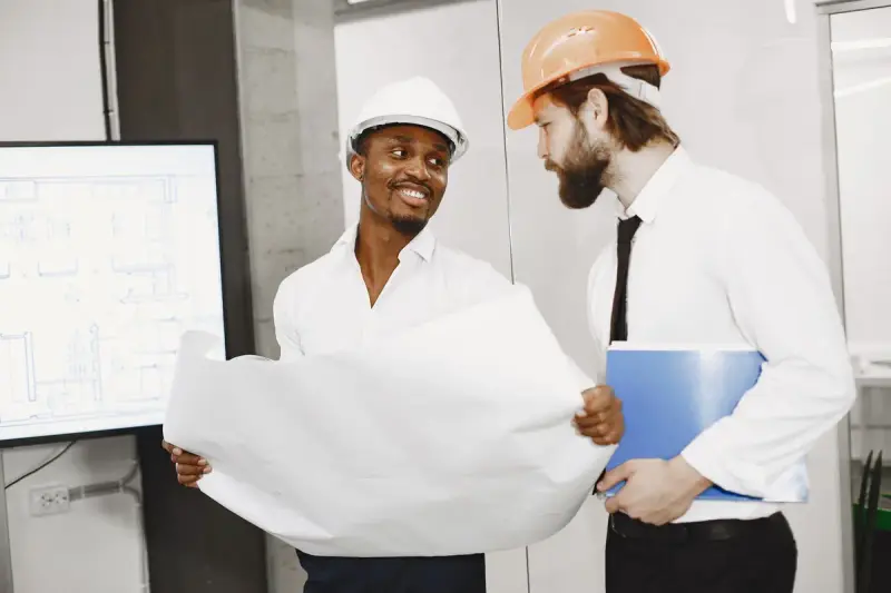 Two architects in hard hats discuss rolled-out plans in a bright office
