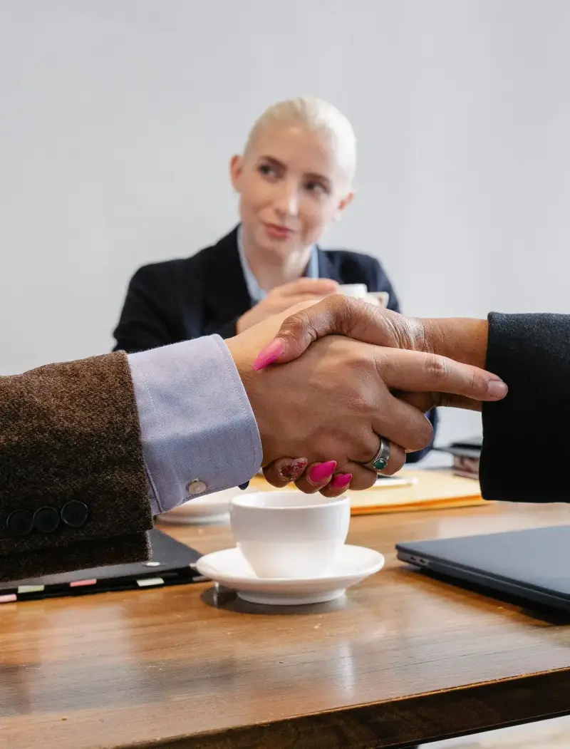 Two colleagues shaking hands across a table with coffee and a laptop