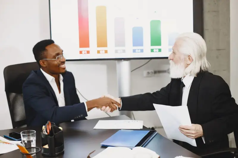 Two businessmen shaking hands at a desk with a bar chart on the wall behind them