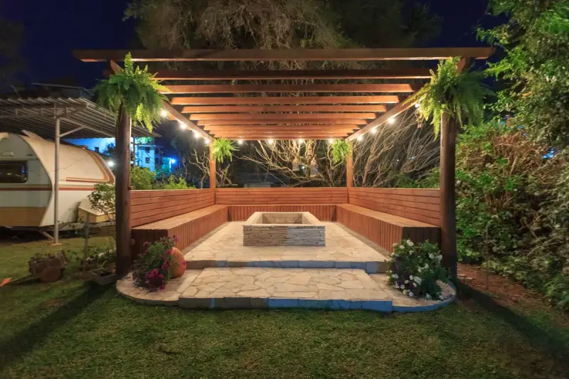 Wooden pergola with string lights over built-in benches and a stone fire pit at night