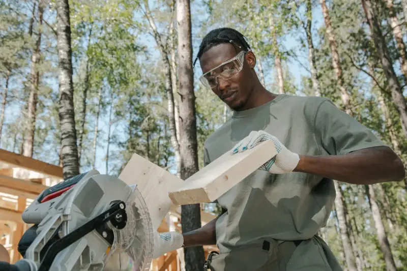 Carpenter in safety glasses and gloves cutting a board on a miter saw among pine trees