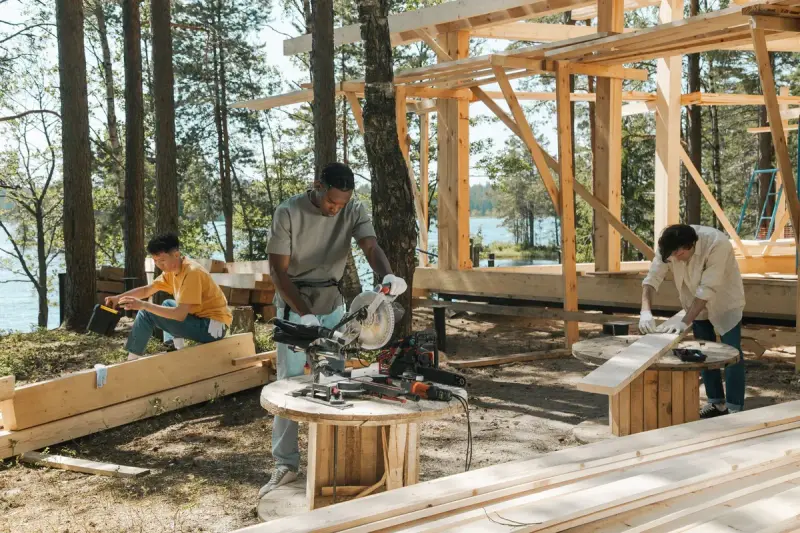 Three carpenters cutting and sanding boards beside a timber frame by a lake