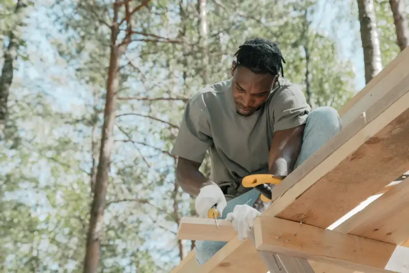 Carpenter in gloves sawing a timber on a wooden roof frame among pine trees