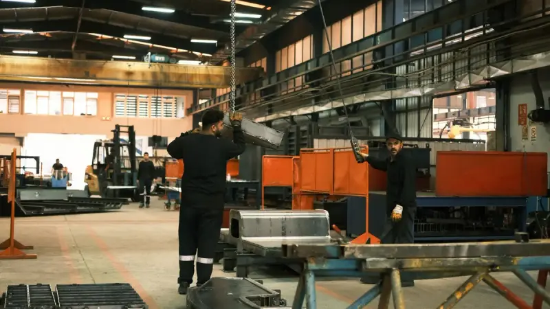Workers at fabrication tables and orange machinery in a long metalworking plant