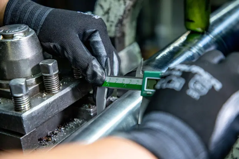 Gloved hands measuring a turned steel bar with a caliper on a lathe
