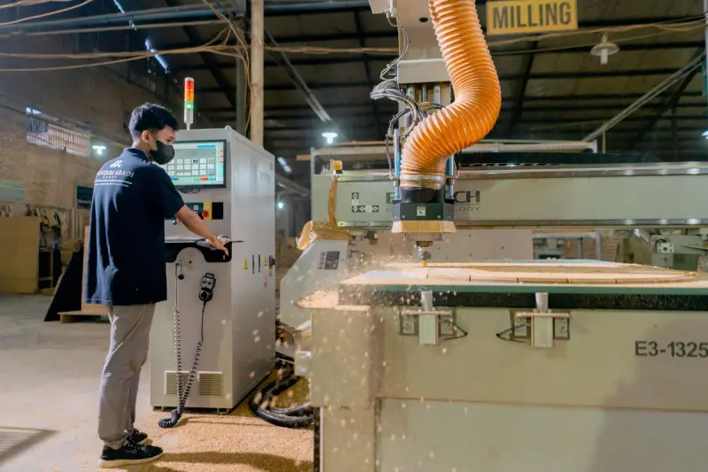 Operator at a control panel while a CNC router cuts panels and chips fly in a busy shop