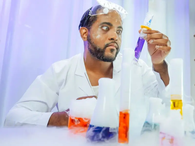 Gloved hand drops blue liquid into a row of test tubes holding colored solutions