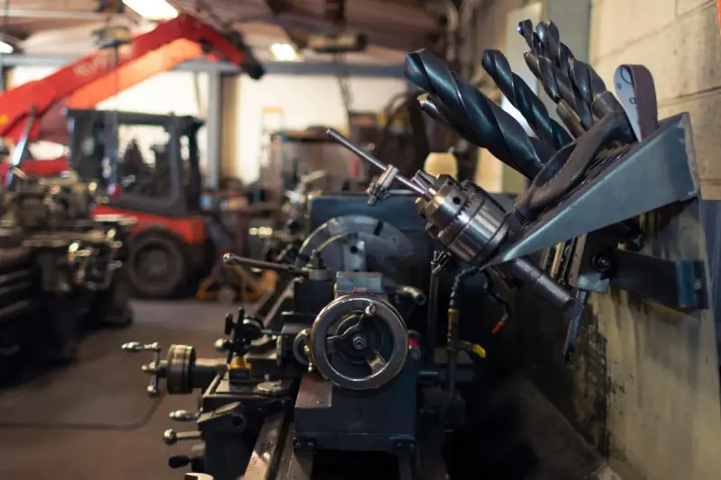 Lathe tool post in front of a rack of large drill bits in a busy machine shop