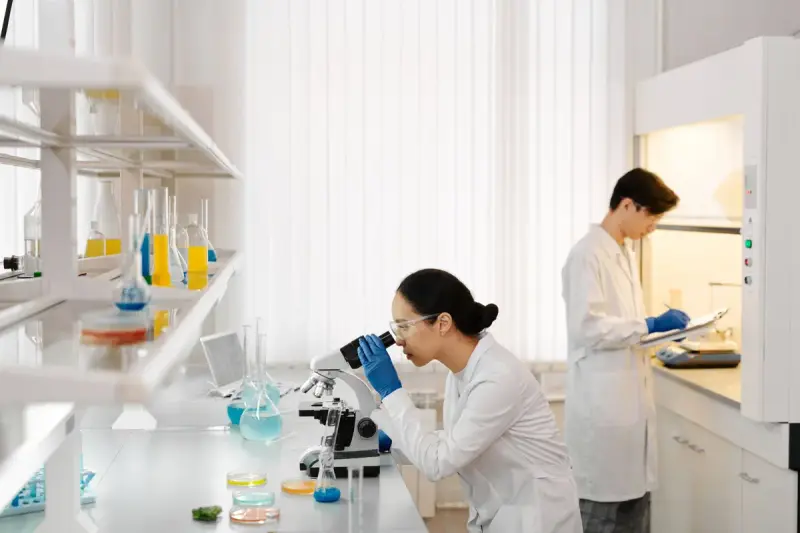 Scientist at a microscope while a colleague records data at a fume hood