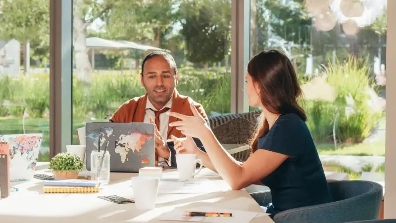 Man and woman in a one-on-one meeting at a table with a laptop by large windows