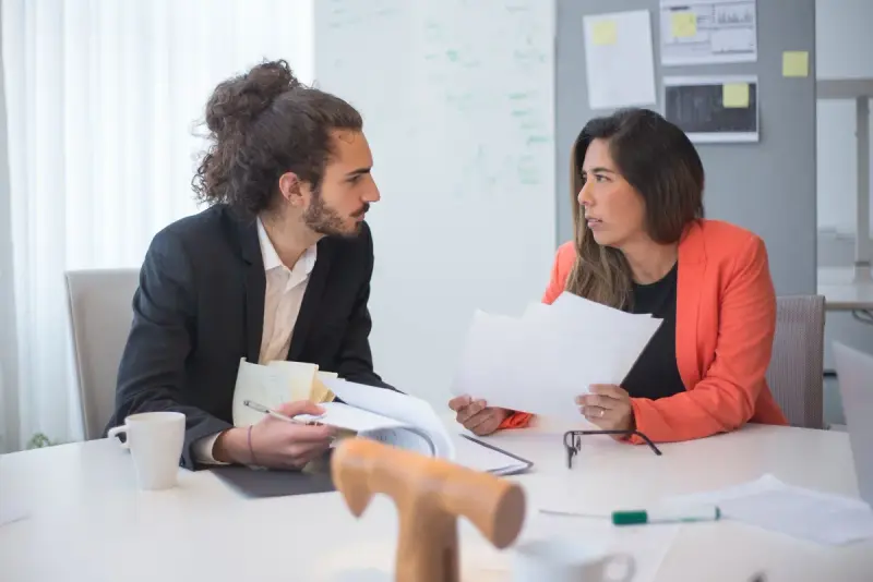 Two colleagues discussing documents across an office table with coffee