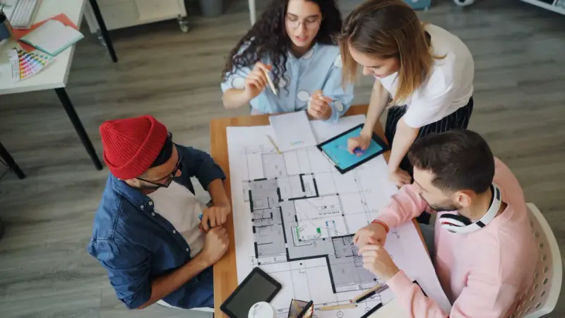Four design professionals marking up a large floor plan around a table with a tablet