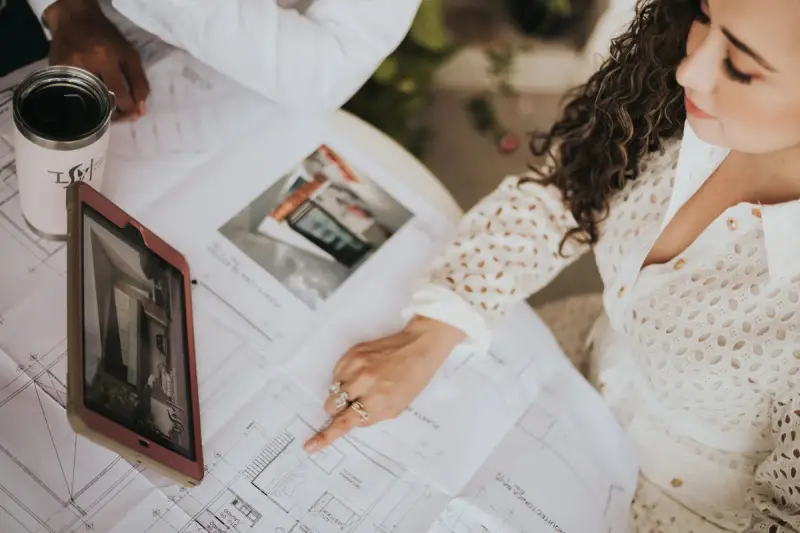 Designer pointing at a layout plan spread on a table beside a tablet and photos