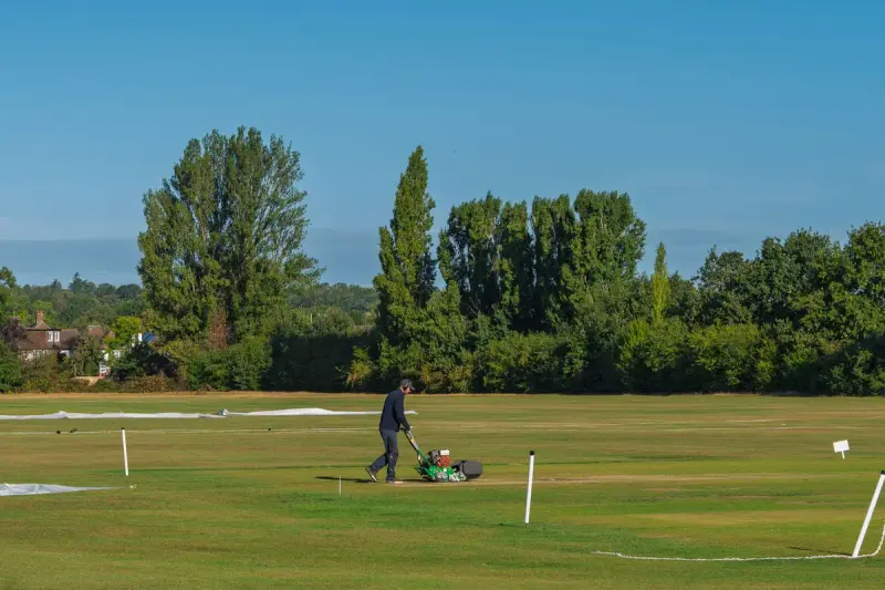 Groundskeeper pushing a mower across a wide sports field lined with tall trees