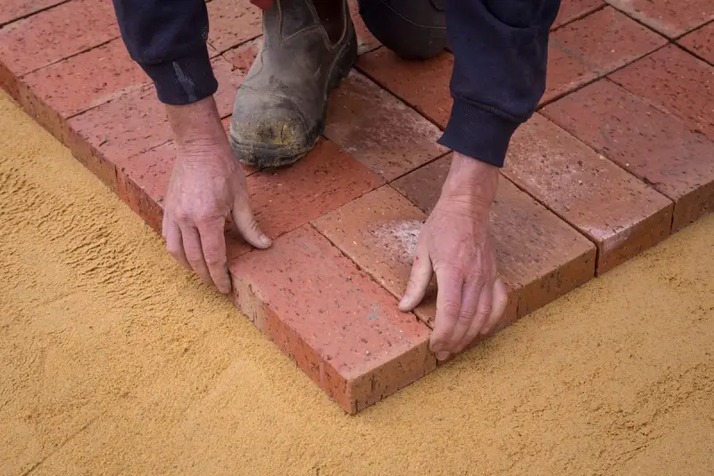Hands setting clay pavers on a bed of sand in a square pattern