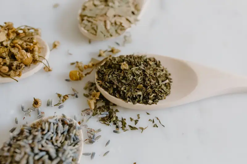 Dried chamomile, lavender and other herbs on wooden spoons on a white marble surface