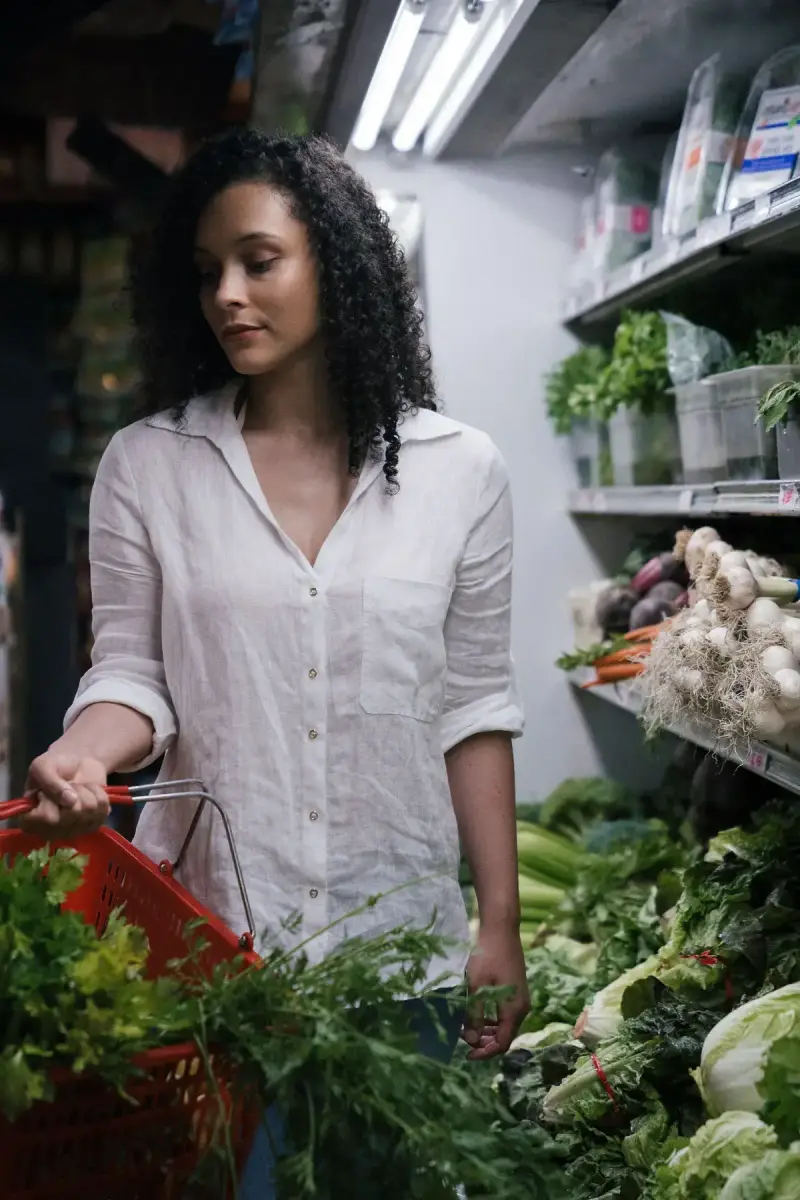 Woman with a red basket of fresh herbs browsing leafy greens in a grocery aisle