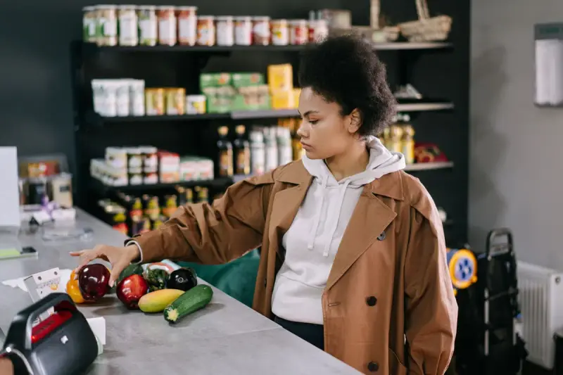 Woman choosing vegetables at a small grocery counter with jars and bottles on shelves behind