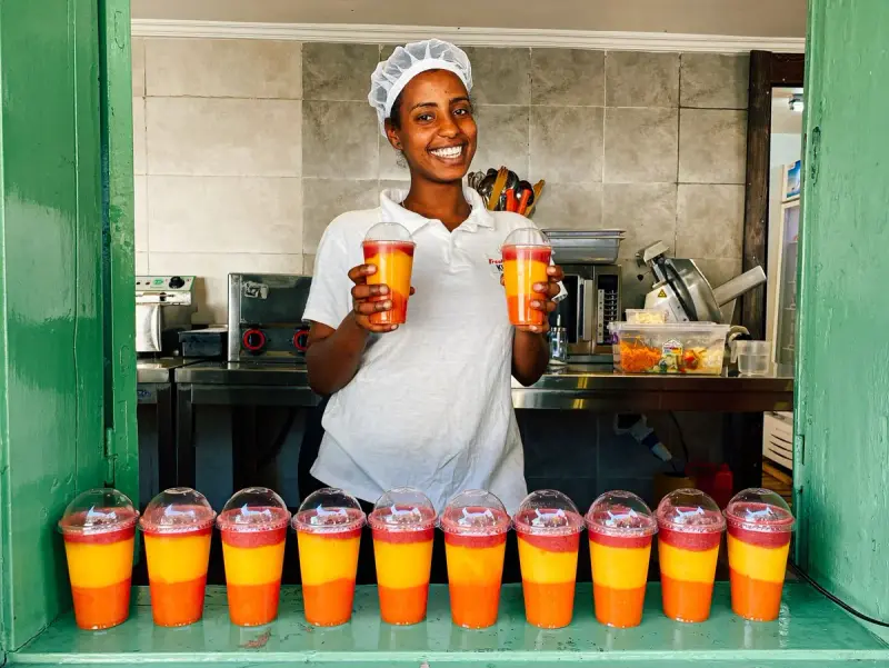 Smiling juice bar worker in a hairnet holds up two layered fruit smoothies above a row of cups