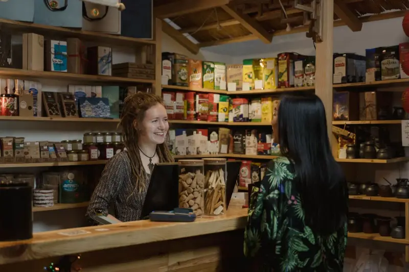 Smiling shopkeeper at a wooden counter helps a customer in a small shop stocked with teas and packaged foods