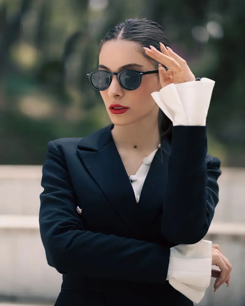 Woman in a black tailored suit touching round dark sunglasses