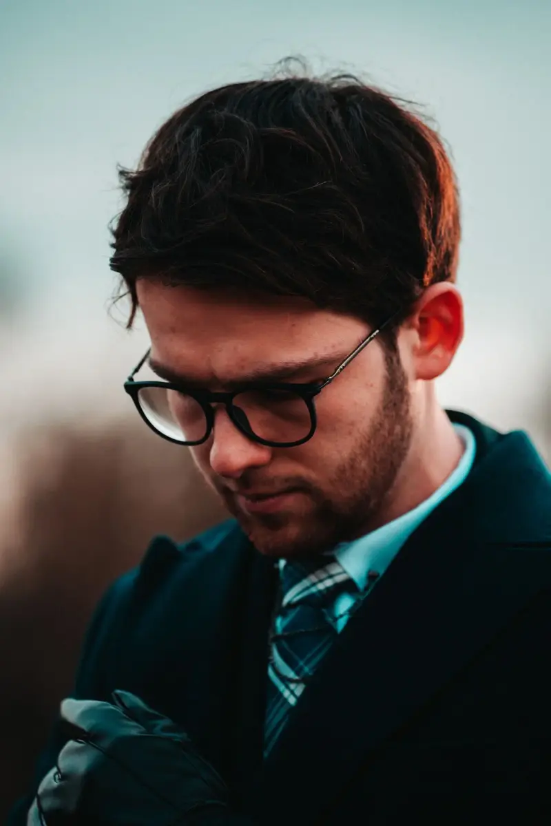Man in a dark coat, tie and black rectangular eyeglasses looking down