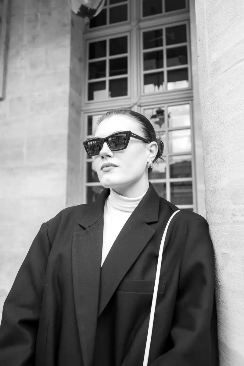 Black and white photo of a woman in cat-eye sunglasses and a long coat by a window