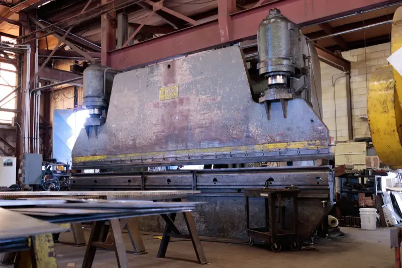 Massive press brake with steel plates stacked on stands in front of it in a working shop