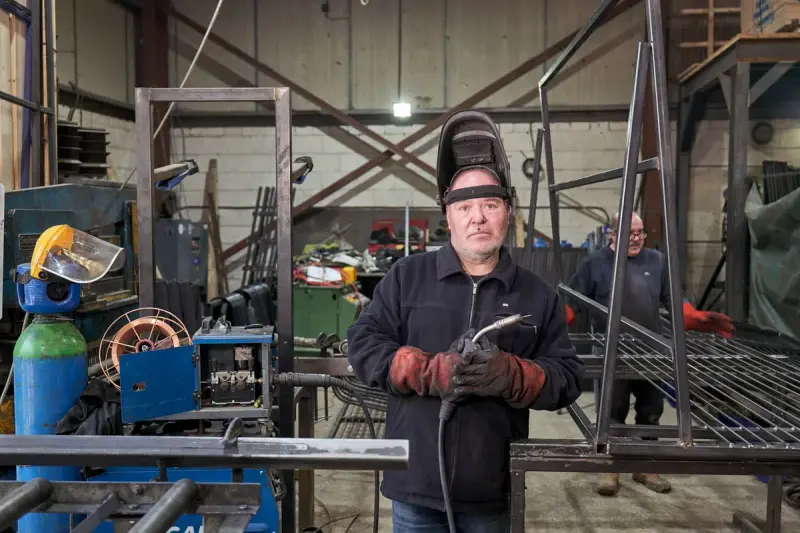 Welder with helmet raised and torch in hand standing in a metal fabrication workshop