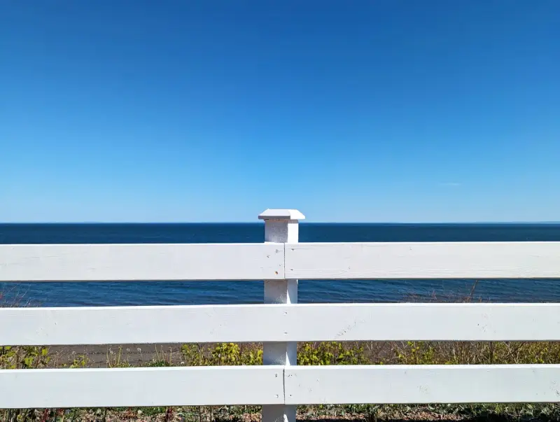 White three-rail wooden fence with a capped post in front of the sea under a clear blue sky
