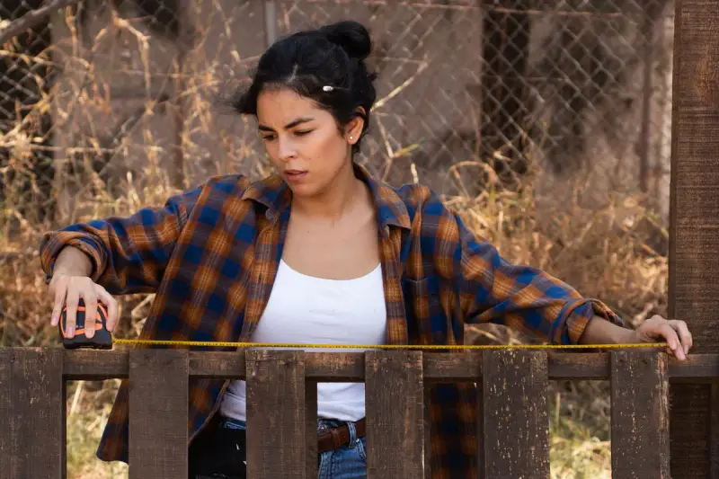 Woman in a flannel shirt measuring a wooden fence rail with a tape measure
