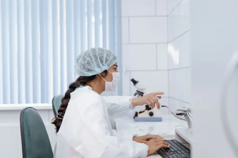 Scientist in a hair net and mask working at a computer beside a microscope