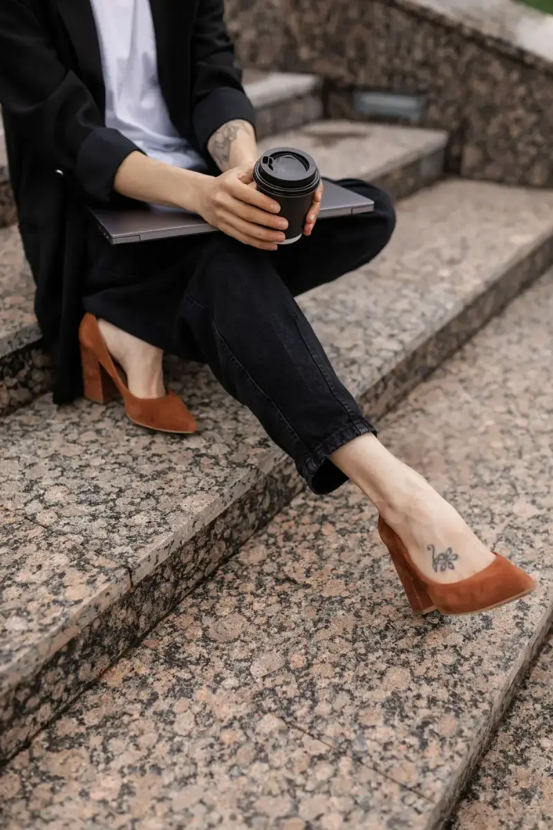 Woman in black trousers and cognac block-heel pumps sitting on granite steps with coffee