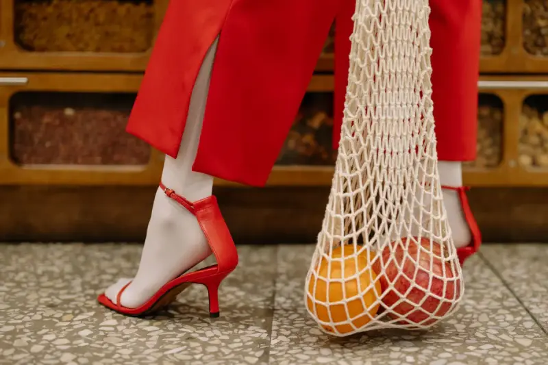 Red strappy heeled sandals and red trousers beside a string bag of oranges