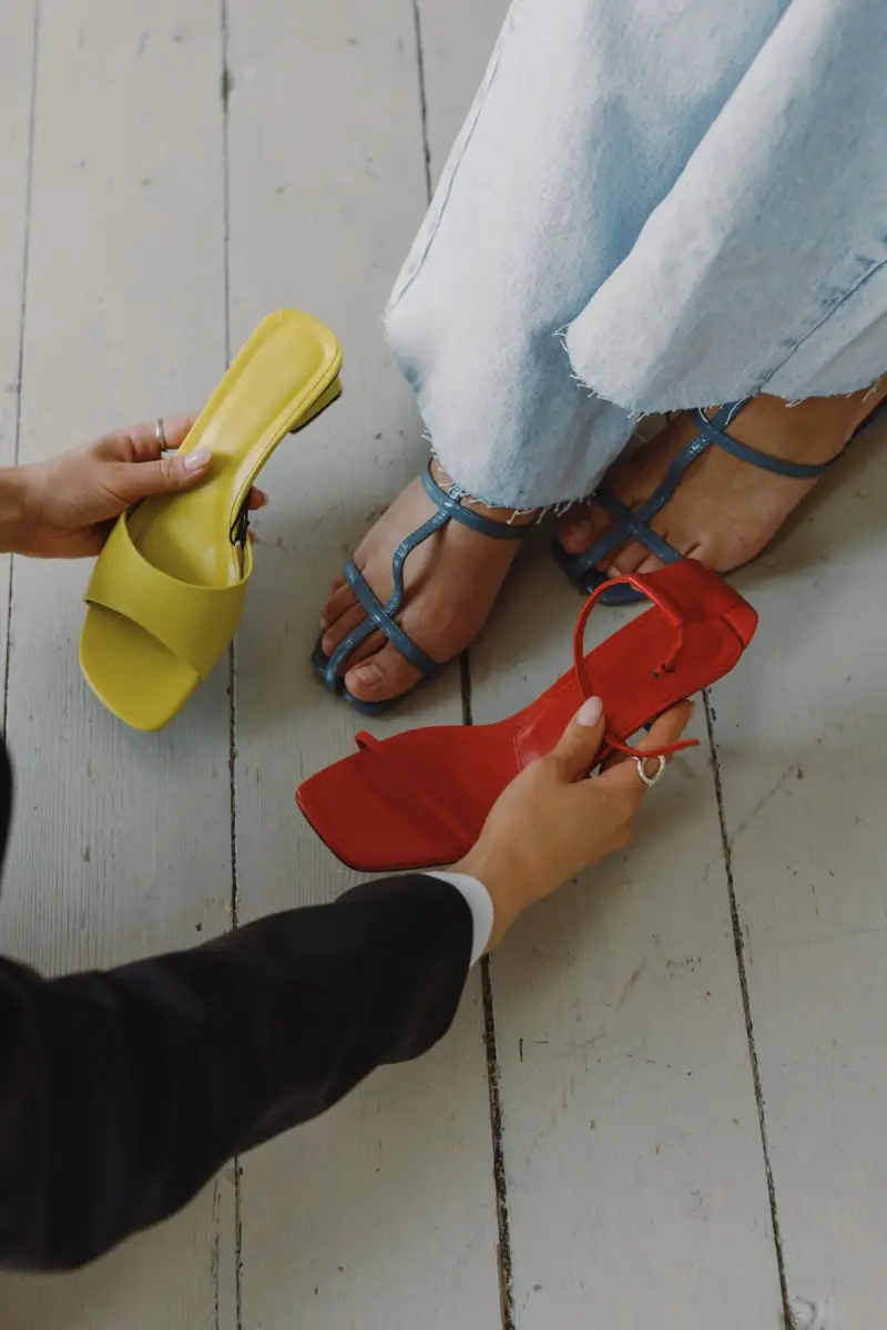 Hands offering yellow and red heeled sandals to a woman trying on shoes in a showroom