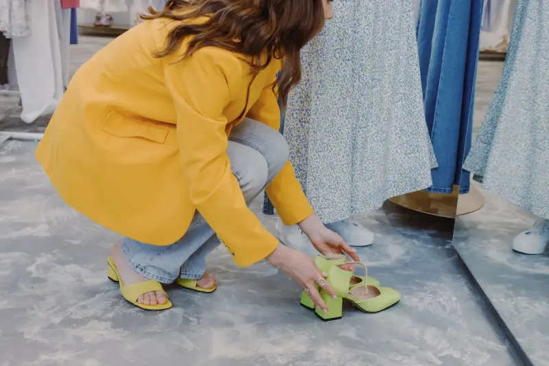 Woman in a yellow blazer crouching to look at green low-heel shoes