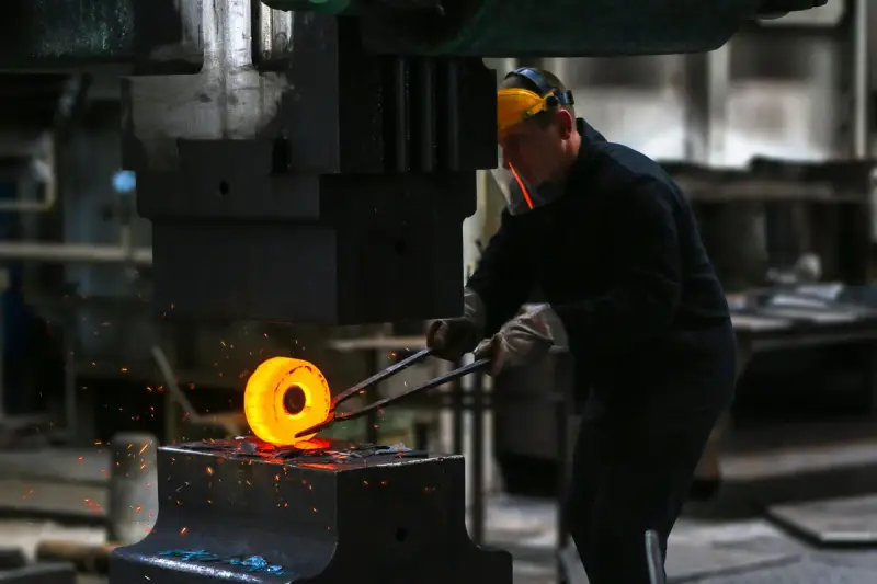 Forge worker in a hard hat holding a glowing steel ring under a forging press
