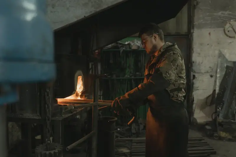 Smith in a leather apron pulling a glowing bar from the furnace with tongs