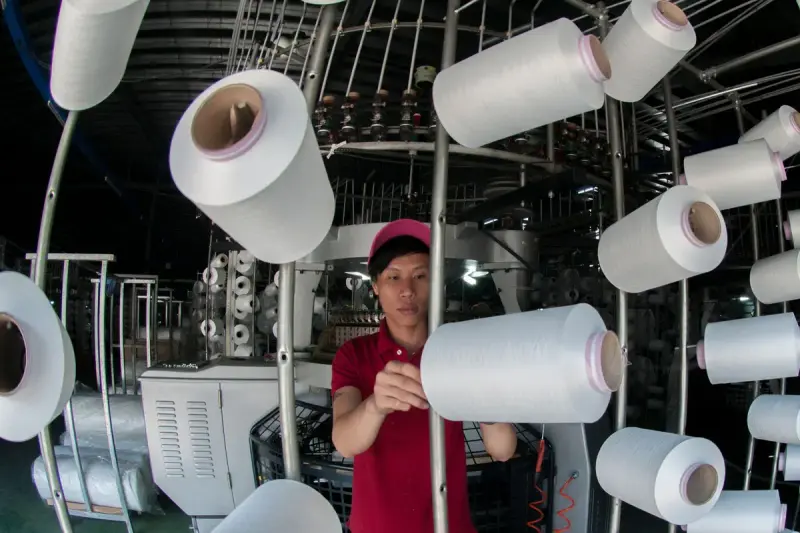 A worker tends yarn spools on a textile machine in a mill