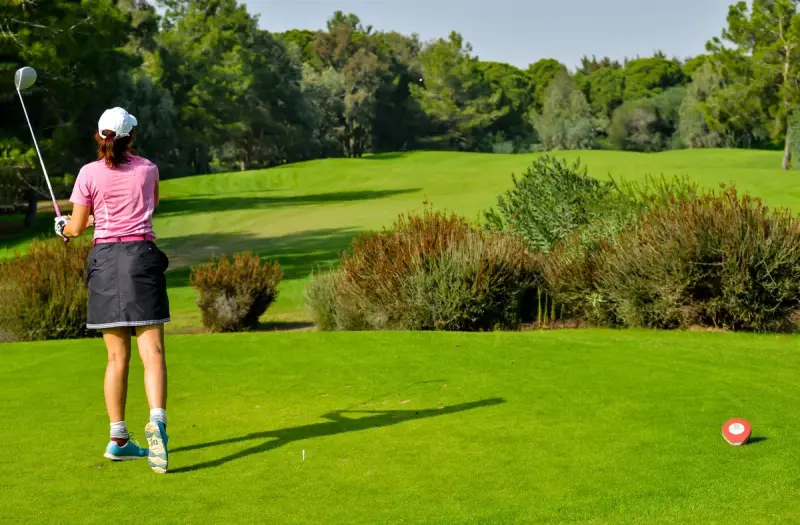 A woman swings a club on a green golf course lined with trees