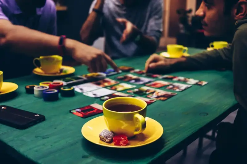 Adults leaning over a card-driven board game on a green table with cups of tea
