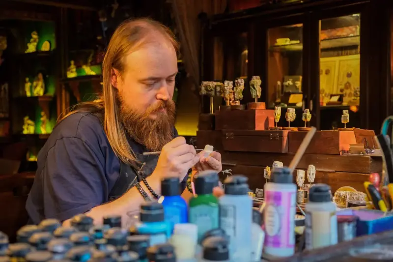 Bearded painter carefully painting a small figure surrounded by paint bottles in a workshop