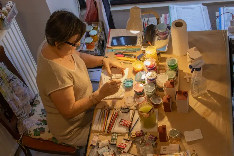 Woman painting small models at a crowded craft table under warm lamplight