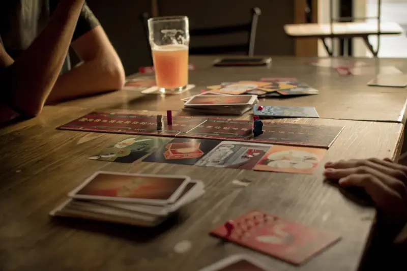 Players around a wooden cafe table covered with game cards and a drink