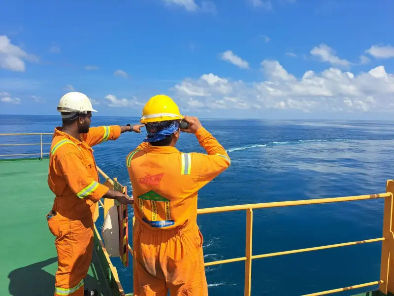 Two workers in orange coveralls and hard hats on a vessel deck looking out to sea