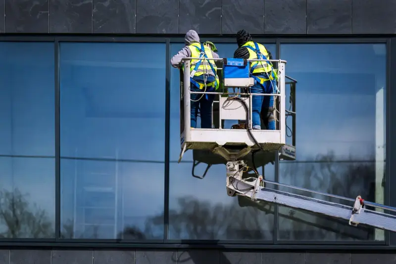 Two workers in safety vests on a boom lift basket working along a dark glass facade