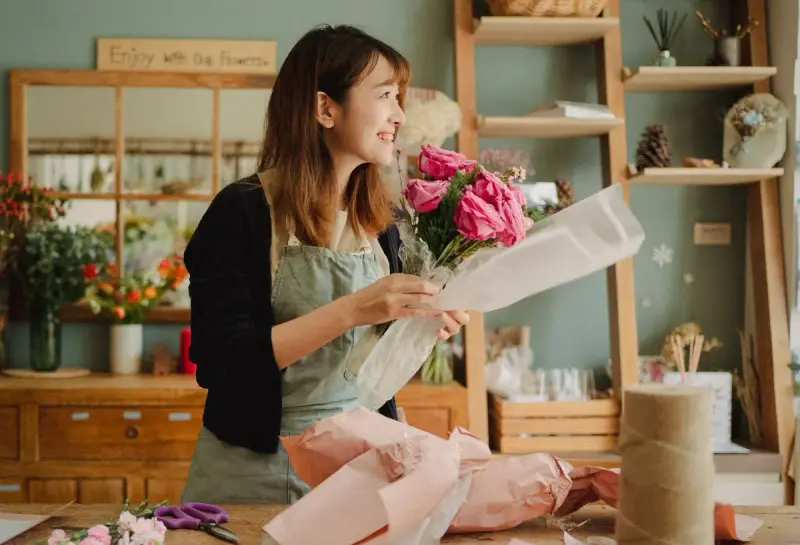 Smiling florist in a green apron wraps a bouquet of pink roses in paper in her flower shop