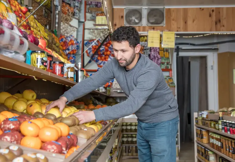 Grocer in a gray sweater arranges oranges, apples and kiwis on a fruit display in a small store