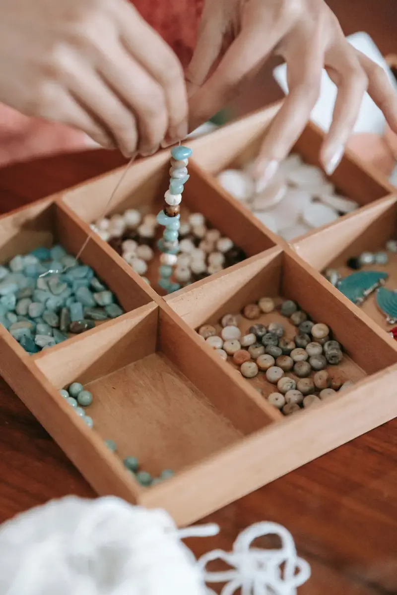 Hands stringing beads on cord over a wooden tray of turquoise and shell beads